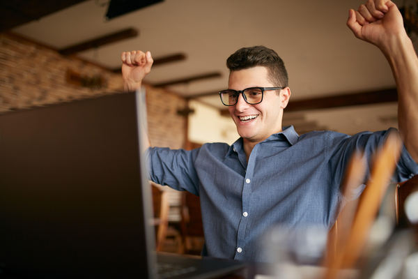 Man sitting in front of his laptop with hands raised in triumph.